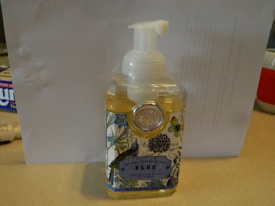 A bottle of liquid hand soap with a pump dispenser on top. The label has floral and butterfly designs and includes text suggesting it's a product from Michel Design Works. The bottle is mostly transparent, revealing the yellowish soap inside. It is standing on a light-colored countertop.