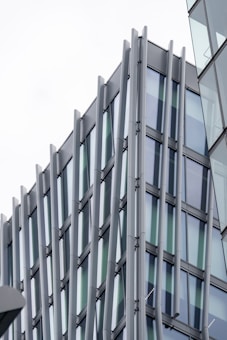 A modern architectural design featuring a building with glass windows and vertical metallic beams. The structure exhibits a sleek and contemporary style with reflective surfaces.