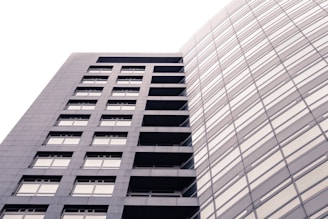 A modern, high-rise building with a sleek, rectangular design and numerous windows. The structure is composed of two distinct sections: one with visible balconies and another with a seamless glass facade. The lighting creates a soft, uniform tone across the building's exterior.