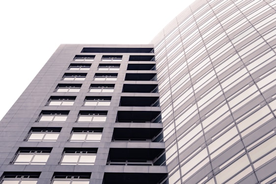 A modern, high-rise building with a sleek, rectangular design and numerous windows. The structure is composed of two distinct sections: one with visible balconies and another with a seamless glass facade. The lighting creates a soft, uniform tone across the building's exterior.
