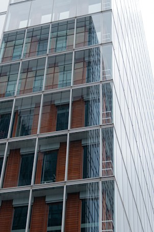 A modern glass skyscraper fa&ccedil;ade featuring large reflective windows, with visible interior elements like wooden paneling. The structure is composed of grid-like patterns with sleek, clean lines.