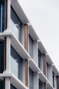 A modern building facade featuring a geometric design with repetitive horizontal and vertical elements. The structure is characterized by textured stone-like surfaces and large glass windows, giving it a contemporary and minimalist aesthetic.
