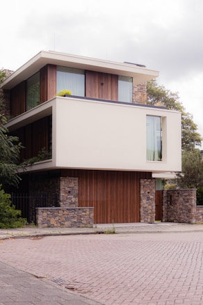 A modern three-story house featuring a blend of stone and wood elements. The building has large glass windows and a flat roof, creating a sleek and contemporary appearance. Lush greenery and trees surround the structure, enhancing the natural look. The street in front is paved with bricks, adding to the clean and tidy environment.