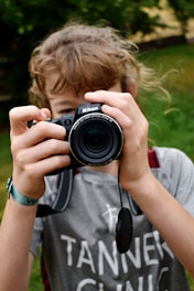 A person with light brown hair holds a Nikon camera up to their face, focusing the lens directly at the viewer. The gray t-shirt features the text 'Tanner Clinic'. The background is blurred greenery, suggesting an outdoor setting.