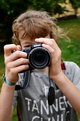 A person with light brown hair holds a Nikon camera up to their face, focusing the lens directly at the viewer. The gray t-shirt features the text 'Tanner Clinic'. The background is blurred greenery, suggesting an outdoor setting.