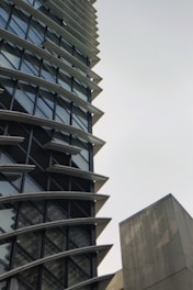 A tall, modern building with a unique architectural design featuring multiple horizontal fins between the windows. The structure has a lot of glass windows, giving it a sleek look. There is also a plain concrete structure visible on the right side.