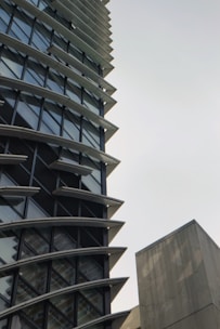 A tall, modern building with a unique architectural design featuring multiple horizontal fins between the windows. The structure has a lot of glass windows, giving it a sleek look. There is also a plain concrete structure visible on the right side.