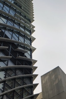A tall, modern building with a unique architectural design featuring multiple horizontal fins between the windows. The structure has a lot of glass windows, giving it a sleek look. There is also a plain concrete structure visible on the right side.