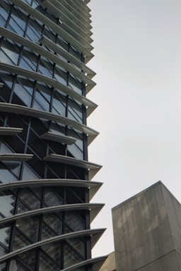 A tall, modern building with a unique architectural design featuring multiple horizontal fins between the windows. The structure has a lot of glass windows, giving it a sleek look. There is also a plain concrete structure visible on the right side.