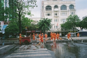 A group of workers wearing orange safety uniforms and helmets are cleaning a paved street area near a crosswalk. They are using hoses and brooms to clean the wet surface. Surrounding buildings, including a hotel named 'ROYAL HOTEL SAIGON,' and trees can be seen in the background.