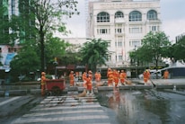 A group of workers wearing orange safety uniforms and helmets are cleaning a paved street area near a crosswalk. They are using hoses and brooms to clean the wet surface. Surrounding buildings, including a hotel named 'ROYAL HOTEL SAIGON,' and trees can be seen in the background.