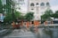 A group of workers wearing orange safety uniforms and helmets are cleaning a paved street area near a crosswalk. They are using hoses and brooms to clean the wet surface. Surrounding buildings, including a hotel named 'ROYAL HOTEL SAIGON,' and trees can be seen in the background.