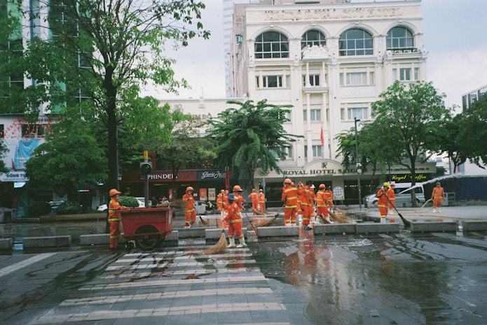 A group of workers wearing orange safety uniforms and helmets are cleaning a paved street area near a crosswalk. They are using hoses and brooms to clean the wet surface. Surrounding buildings, including a hotel named 'ROYAL HOTEL SAIGON,' and trees can be seen in the background.