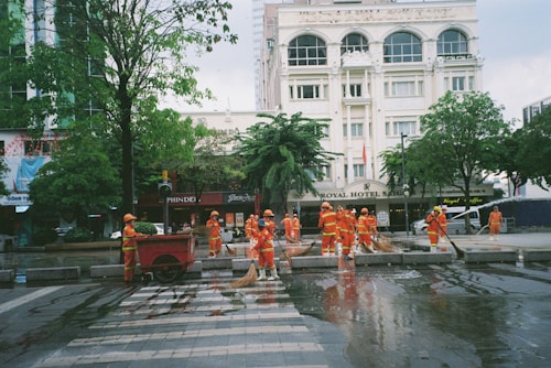 A group of workers wearing orange safety uniforms and helmets are cleaning a paved street area near a crosswalk. They are using hoses and brooms to clean the wet surface. Surrounding buildings, including a hotel named 'ROYAL HOTEL SAIGON,' and trees can be seen in the background.