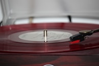 A close-up view of a record player turntable with a red vinyl record spinning on it. The tonearm and needle are positioned on the record, suggesting that music is being played. The background is blurred, focusing attention on the record and player.