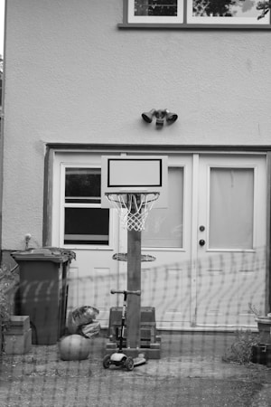 A black and white image of a backyard scene featuring a basketball hoop mounted on a pole. In front of the hoop, there is a scooter and a basketball resting on the ground. A trash bin is positioned to the left, and there are bricks stacked near the base of the pole. The backdrop consists of double doors with frosted windows attached to a house.