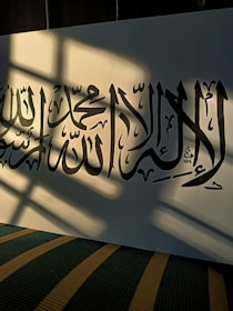 Arabic calligraphy is elegantly written on a sheet of paper, with sunlight casting shadows across the page, creating a dramatic contrast. The text is black on a white background, and the scene conveys a serene atmosphere.