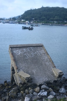 A concrete structure with the words 'DON'T JUMP' written on it in white chalk stands on rocky terrain near a body of water. In the background, there are boats on the water and a wooded hilly landscape with a few buildings and scattered structures.