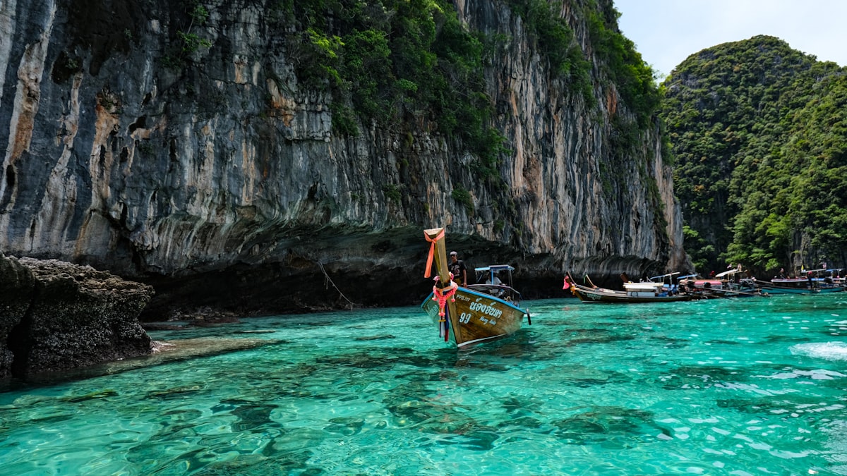 Several colorful long-tail boats with curved prows float on calm emerald green water beneath dramatic limestone cliffs rising from the sea.. Photo by Ashiks Visual on Unsplash.