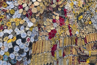 A complex and vibrant collage composed of an array of colorful bottle caps and metal pieces. The elements include bright yellows, deep reds, various shades of gold, and some blues and whites. The materials are intricately layered and connected, creating a textured and visually dynamic surface. The bottle caps include recognizable logos and text, adding a pop-culture and recycled art element to the piece.
