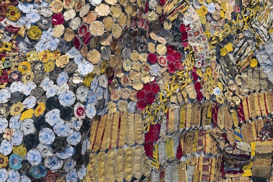 A complex and vibrant collage composed of an array of colorful bottle caps and metal pieces. The elements include bright yellows, deep reds, various shades of gold, and some blues and whites. The materials are intricately layered and connected, creating a textured and visually dynamic surface. The bottle caps include recognizable logos and text, adding a pop-culture and recycled art element to the piece.