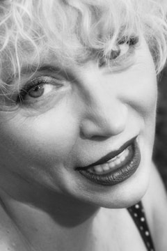 Close-up of a person's face with curly blonde hair and a warm smile. The image has a vintage black and white effect, highlighting the eyes and texture of the skin.