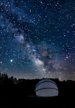 A night sky filled with numerous stars and the Milky Way galaxy prominently visible across the expanse. There is a white observatory dome in the foreground, partially illuminated, situated among dark silhouettes of trees.