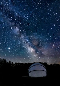 A night sky filled with numerous stars and the Milky Way galaxy prominently visible across the expanse. There is a white observatory dome in the foreground, partially illuminated, situated among dark silhouettes of trees.