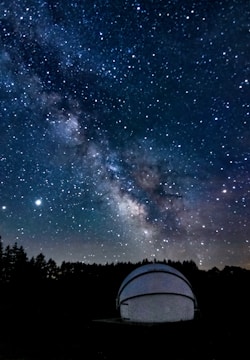 A night sky filled with numerous stars and the Milky Way galaxy prominently visible across the expanse. There is a white observatory dome in the foreground, partially illuminated, situated among dark silhouettes of trees.