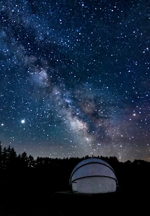 A night sky filled with numerous stars and the Milky Way galaxy prominently visible across the expanse. There is a white observatory dome in the foreground, partially illuminated, situated among dark silhouettes of trees.