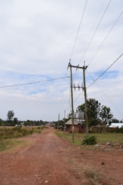 A rural dirt road stretches into the distance with power lines running parallel on wooden poles. There are scattered houses and vegetation on either side, with a cloudy sky overhead.