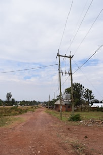 A rural dirt road stretches into the distance with power lines running parallel on wooden poles. There are scattered houses and vegetation on either side, with a cloudy sky overhead.