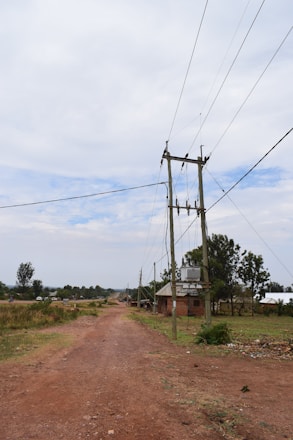 A rural dirt road stretches into the distance with power lines running parallel on wooden poles. There are scattered houses and vegetation on either side, with a cloudy sky overhead.