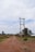 A rural dirt road stretches into the distance with power lines running parallel on wooden poles. There are scattered houses and vegetation on either side, with a cloudy sky overhead.