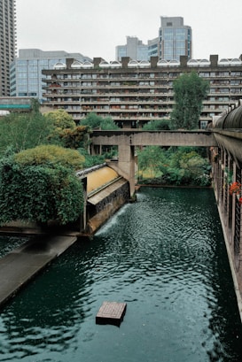 A modern urban setting featuring a large residential building with multiple balconies and greenery. The foreground shows a water feature with clear, rippling water and a small platform. Surrounding the water is lush, well-maintained vegetation. In the distance, tall glass and concrete buildings rise against the sky.