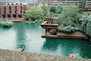 An urban landscape featuring a modern architectural style with a series of buildings and a prominent water body. Rich greenery surrounds the structures, adding natural elements to the concrete environment. The water has a still, teal appearance, reflecting some of the surrounding greenery and buildings.