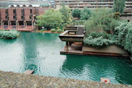 An urban landscape featuring a modern architectural style with a series of buildings and a prominent water body. Rich greenery surrounds the structures, adding natural elements to the concrete environment. The water has a still, teal appearance, reflecting some of the surrounding greenery and buildings.