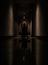 A person is sitting cross-legged on the floor in a dark corridor. Their hands are raised and joined above their head, creating a silhouette effect. A strong overhead light casts a reflection on the polished floor, enhancing the symmetry of the scene.