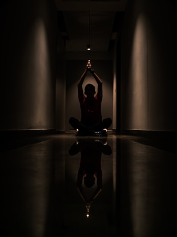 A person is sitting cross-legged on the floor in a dark corridor. Their hands are raised and joined above their head, creating a silhouette effect. A strong overhead light casts a reflection on the polished floor, enhancing the symmetry of the scene.