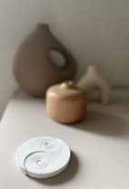 A minimalist composition featuring three ceramic objects in neutral tones, arranged on a flat surface. A white textured circular object with a yin-yang pattern is prominently placed in the foreground, while blurred background objects include vase-like forms in earth tones.