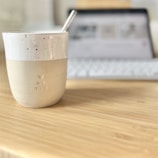 A ceramic cup with a speckled design and the words 'ME TIME' embossed on it sits on a wooden table. The cup has a straw inside, and in the background, a blurred-out laptop with a keyboard is visible, suggesting a cozy workspace setup.