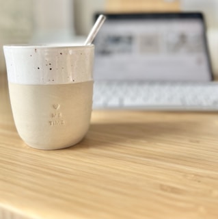 A ceramic cup with a speckled design and the words 'ME TIME' embossed on it sits on a wooden table. The cup has a straw inside, and in the background, a blurred-out laptop with a keyboard is visible, suggesting a cozy workspace setup.