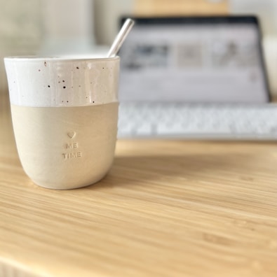 A ceramic cup with a speckled design and the words 'ME TIME' embossed on it sits on a wooden table. The cup has a straw inside, and in the background, a blurred-out laptop with a keyboard is visible, suggesting a cozy workspace setup.