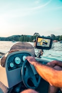 A steering wheel and dashboard of a boat with various gauges is visible. A person is operating the steering wheel while next to it, an electronic display shows navigation data. In the background, a serene body of water with boats and distant lush greenery can be seen under a clear sky.