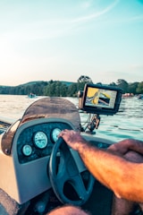 A steering wheel and dashboard of a boat with various gauges is visible. A person is operating the steering wheel while next to it, an electronic display shows navigation data. In the background, a serene body of water with boats and distant lush greenery can be seen under a clear sky.