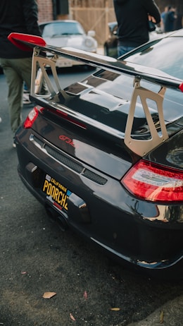 A close-up of a black car with a prominent rear spoiler, displaying a custom California license plate reading 'POORCH'. The taillights are prominent, and the setting seems to be an outdoor or street scene with other vehicles and people partially visible in the background.