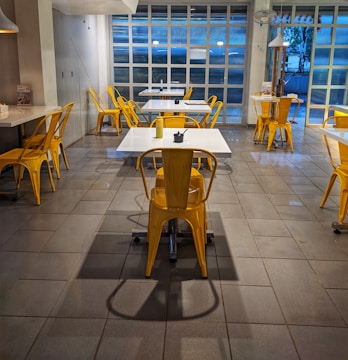 An indoor dining area features sleek, modern design with yellow metal chairs and white tables arranged in a spacious layout. Bright natural light streams through large grid-patterned windows, creating a welcoming ambiance. The floor is tiled in stone gray, reflecting a clean and contemporary style.