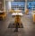 An indoor dining area features sleek, modern design with yellow metal chairs and white tables arranged in a spacious layout. Bright natural light streams through large grid-patterned windows, creating a welcoming ambiance. The floor is tiled in stone gray, reflecting a clean and contemporary style.