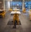 An indoor dining area features sleek, modern design with yellow metal chairs and white tables arranged in a spacious layout. Bright natural light streams through large grid-patterned windows, creating a welcoming ambiance. The floor is tiled in stone gray, reflecting a clean and contemporary style.