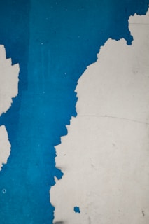 Peeling blue paint on a rough surface, revealing an underlying layer. The jagged lines of the chipped paint create an abstract and textured pattern, with stark contrast between the vibrant blue and the exposed lighter layer.